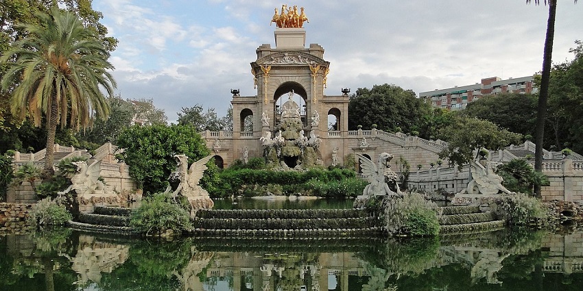 Elegant stone fountain with statues, greenery, and a golden sculpture on an arched structure.