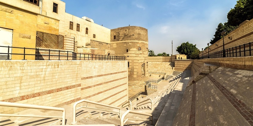 Ancient fortress tower with modern stone amphitheatre steps, bathed in golden sunlight.