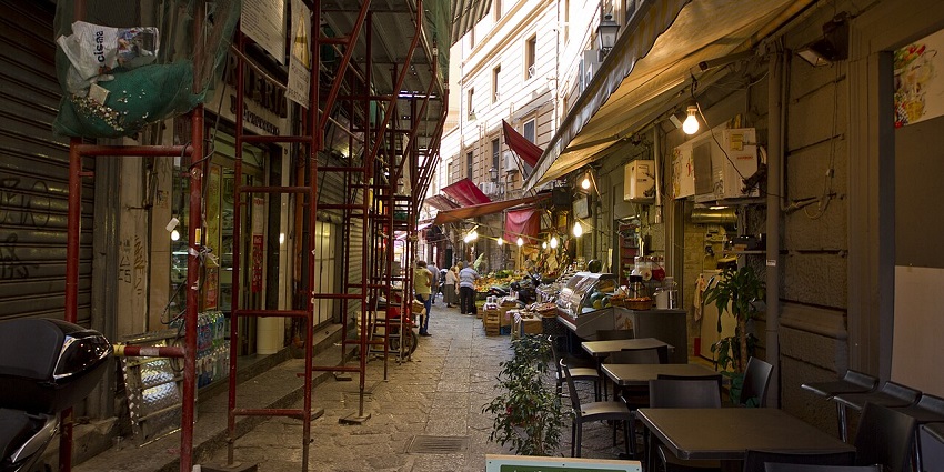 Historic Vucciria market in Palermo with multiple shops and cafes, chairs on the street.
