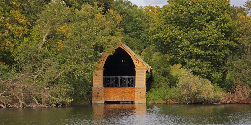 Wooden boathouse with an arched roof surrounded by dense green trees by a calm river.
