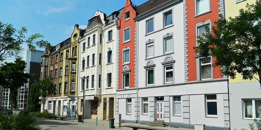 Colourful row of residential buildings with facades lining a quiet, tree-shaded plaza.