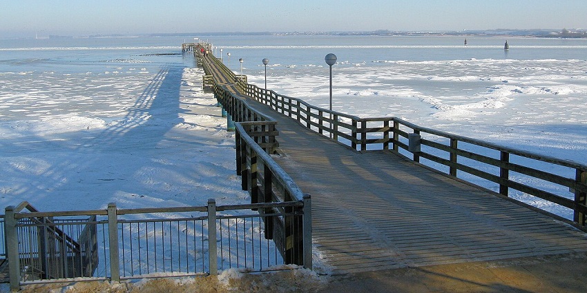 A wooden pier stretches across a frozen sea under a clear blue sky on a crisp winter day.
