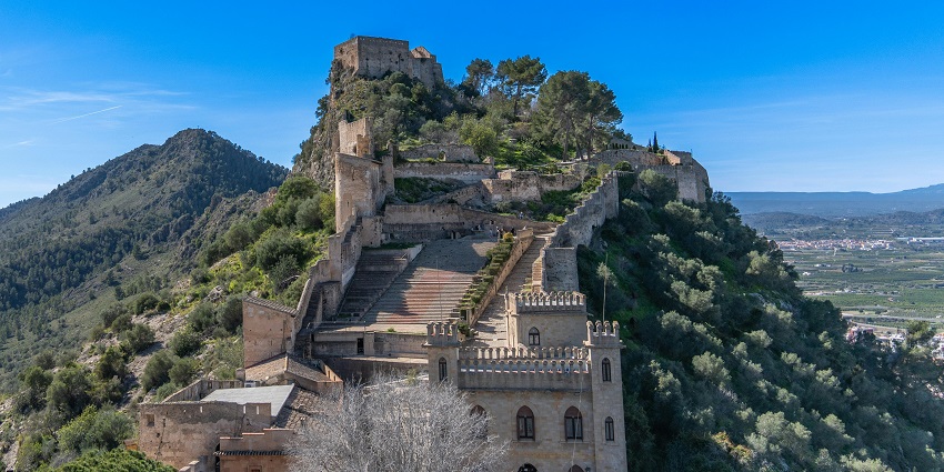 Stone towers and ancient walls of Xàtiva Castle surrounded by green hills and valleys.