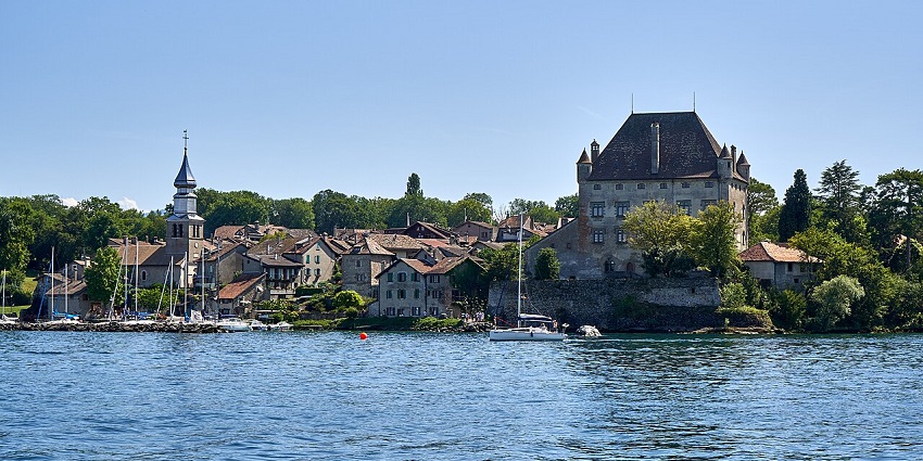 Charming lakeside village with stone buildings, church tower, and sailboats on calm water, amongst the best places to visit near Geneva.