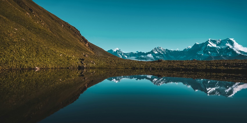 Calm mountain lake in Auli surrounded by rocky slopes and distant snowy peaks.

