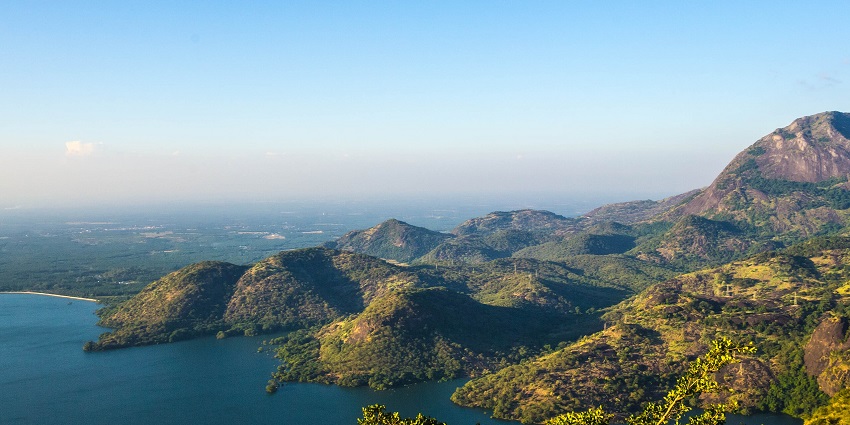 Aliyar Dam Reservoir is surrounded by hills and greenery under a clear blue sky.