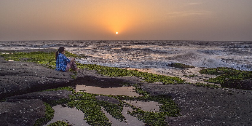 A woman sitting on mossy rocks watching the ocean waves and sunset over the horizon.