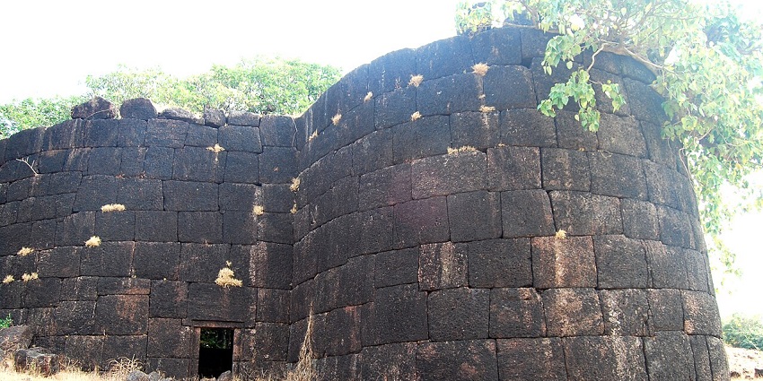Ancient stone fort wall with small entrance, surrounded by dry grass and green foliage.