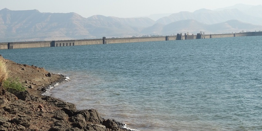 Bhatghar Dam with blue water, muddy shores, and distant dam structure, one of the places to visit near Baneshwar Temple.