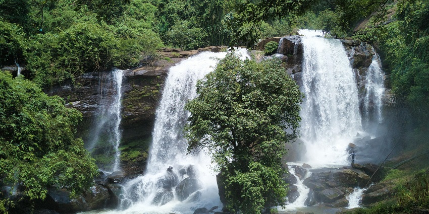 Bhatta Falls, with water cascading over rocky cliffs, is  surrounded by lush greenery.
