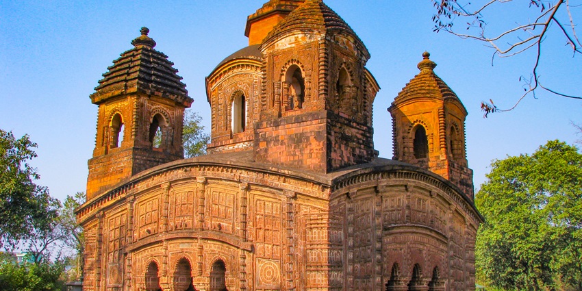 A terracotta temple with ornate carvings and bent towers with trees in the background.