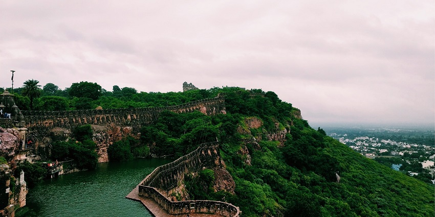 Old fort walls winding over a lush green hill next to a large green water reservoir.
