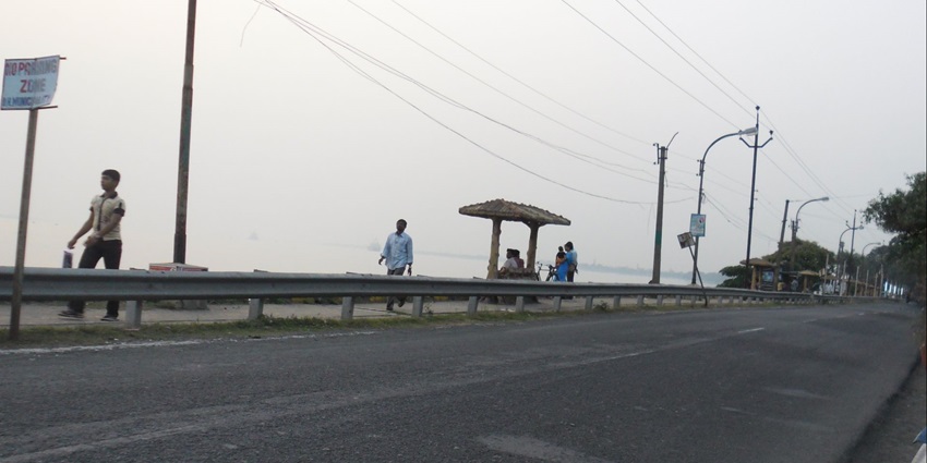 A riverside road with lamp posts, barriers, and huts facing a calm, hazy water view
