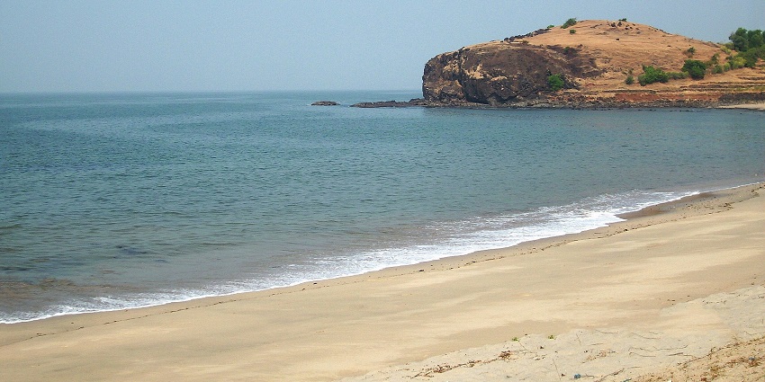 A quiet beach with gentle waves, a rocky hill in the background, and a calm shoreline.
