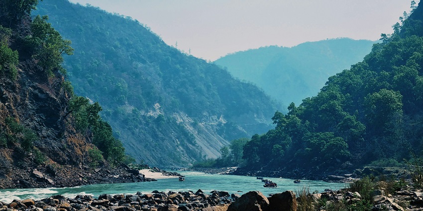 A serene view of the river with a calm water body surrounded by dense greenery and mountains.