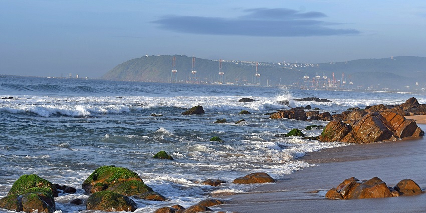 Waves crash on a sandy beach with mossy rocks and distant hills under a soft blue sky, one of the best places to visit near Vizag for 3 days.
