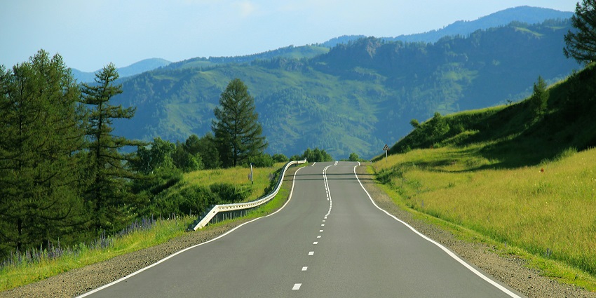 Road winding through lush green hills with distant mountains under clear sky.
