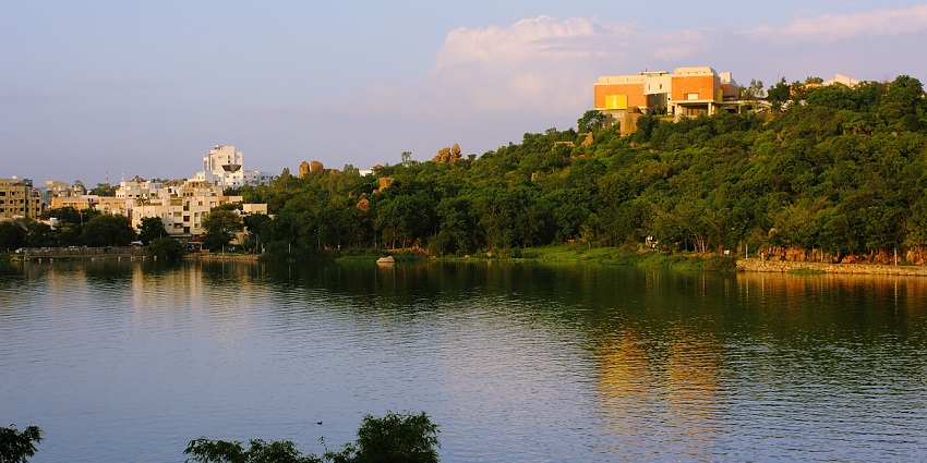 Durgam Cheruvu is a lake reflecting greenery and some distant buildings against a clear blue sky, one of the best places to visit near Gachibowli.