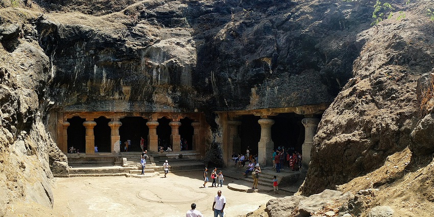 Ancient rock-cut cave entrance with pillars and visitors standing at the wide opening.