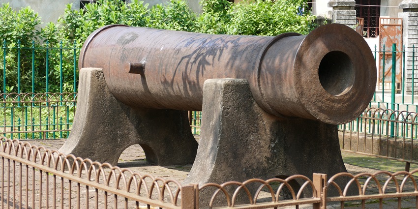 A historic cannon is displayed on a stone platform behind a fence in a shaded courtyard.