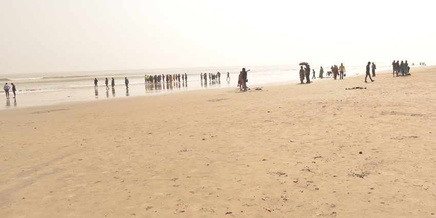People strolling and gathering along a wide sandy beach under a bright, afternoon sky.
