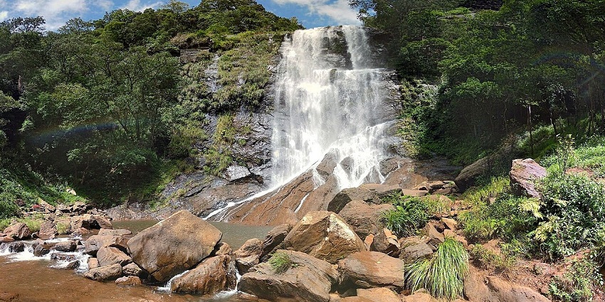 Wide waterfall flowing over rocks into a stream with surrounding greenery and boulders