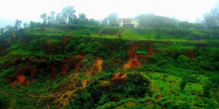 Cliffside view with red rocky slopes, vegetation, and a house on top in misty weather