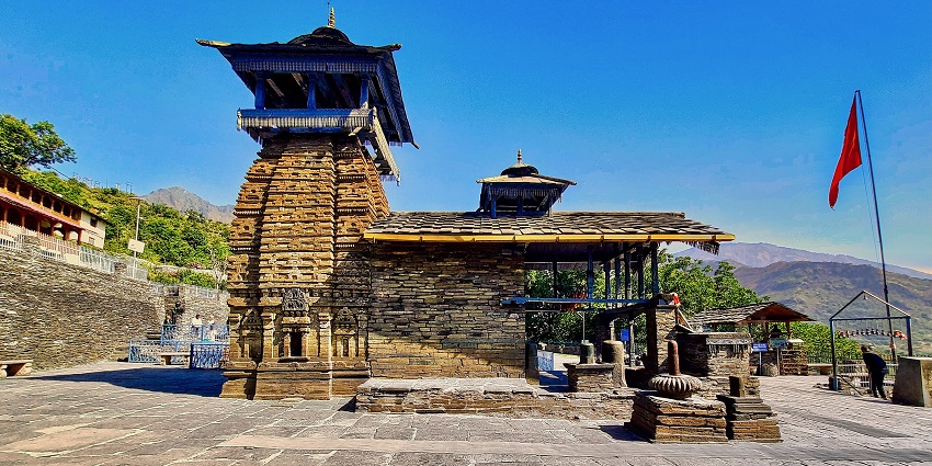 Ancient stone temple with wooden roof and red flag, set against a bright blue sky.
