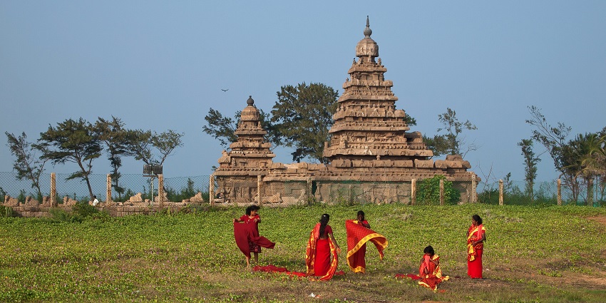 A group of playful women wearing red sarees stand on a field near a temple in Mahabalipuram.