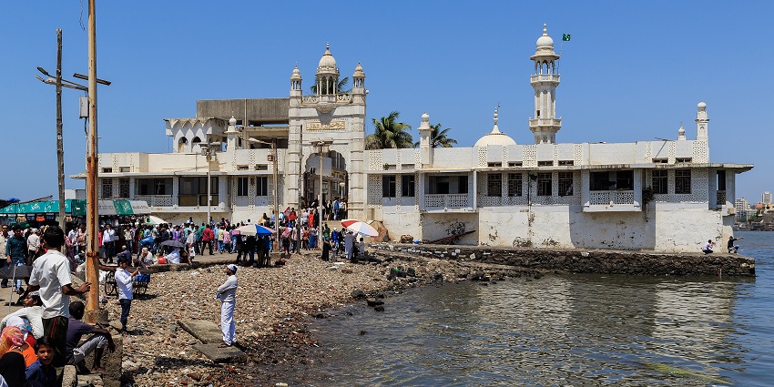 White seaside structure with domes and minarets, crowded with visitors along the shore.
