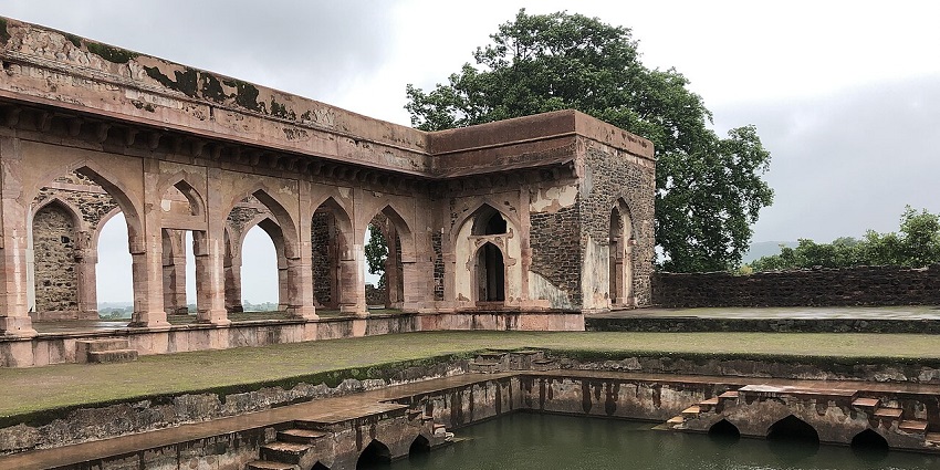 An ancient stone structure with arches surrounding a water tank under a cloudy sky, one of the places to visit near Indore in July.

