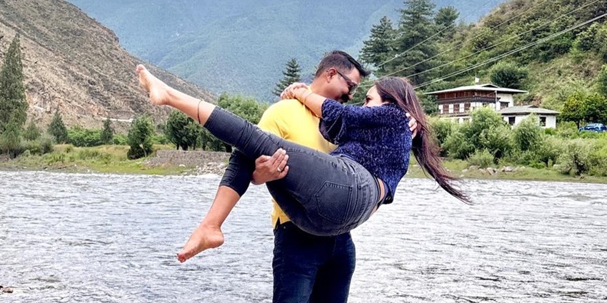 Man lifts woman by a riverbank with green hills and Bhutanese houses in background.