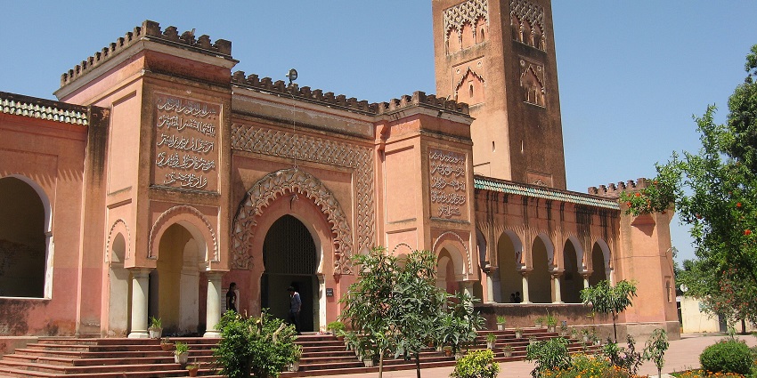 Historic red sandstone building with arched doorways, a tall minaret, and a garden in front.
