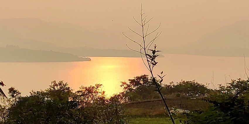 Orange sunset reflecting on calm lake water, framed by silhouetted plants and hills, one of the best places to visit near Pune in monsoon for couples.