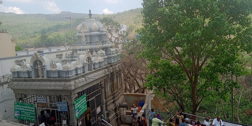 Devotees gather outside a grey temple gateway under a large tree and cloudy mountain sky, one of the best places to visit near Tirupati in one day.