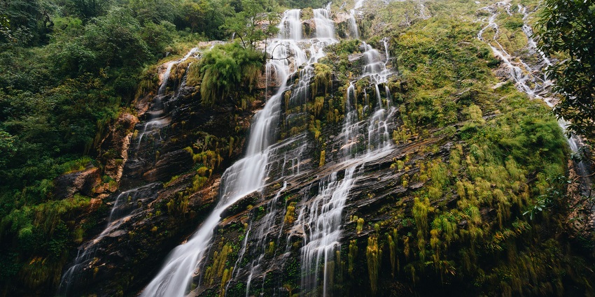 Waterfall streams down rocky cliffs with green hills and a cloudy sky in the background.
