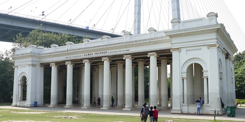 Prinsep Ghat in Kolkata with traditional English architecture and visitors by the river.
