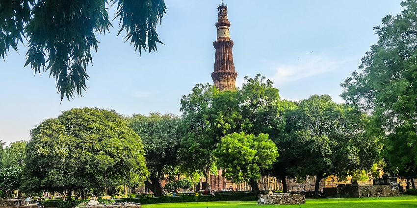 Qutub Minar, visible from afar during the day with a blue sky and green trees all around.