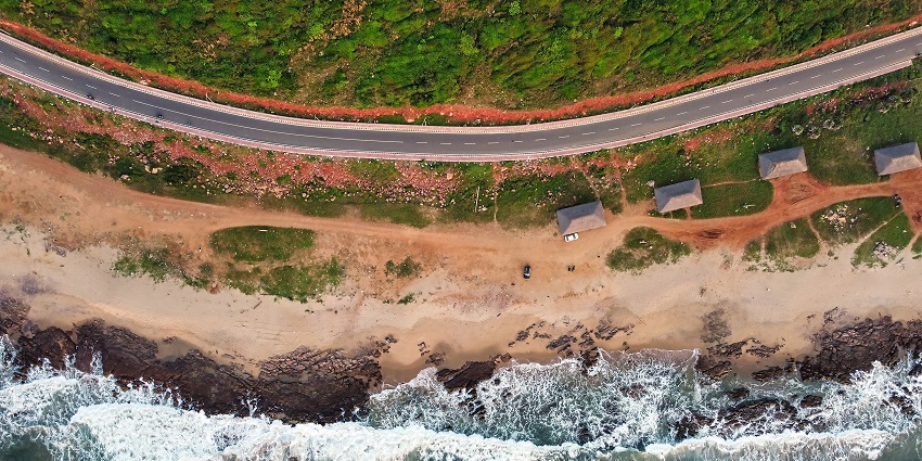 Curving coastal road lined with palm trees beside sandy beach and rolling blue ocean waves, one of the best places to visit near Vizag for 3 days.