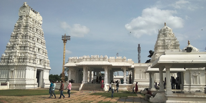 White gopuram towering over landscaped gardens with people walking under a blue sky.
