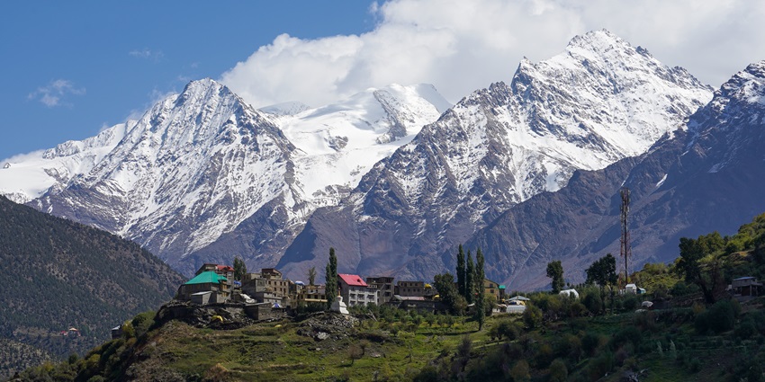 Snow-capped peaks towering over a hillside village with green fields and tall trees