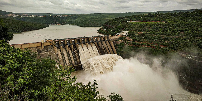 Wide view of Singda Dam surrounded by green hills under a bright, cloudy sky.
