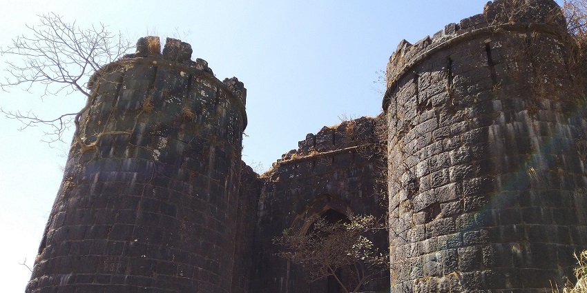 Ancient stone building with intricate carvings surrounded by dry plants under clear blue sky.