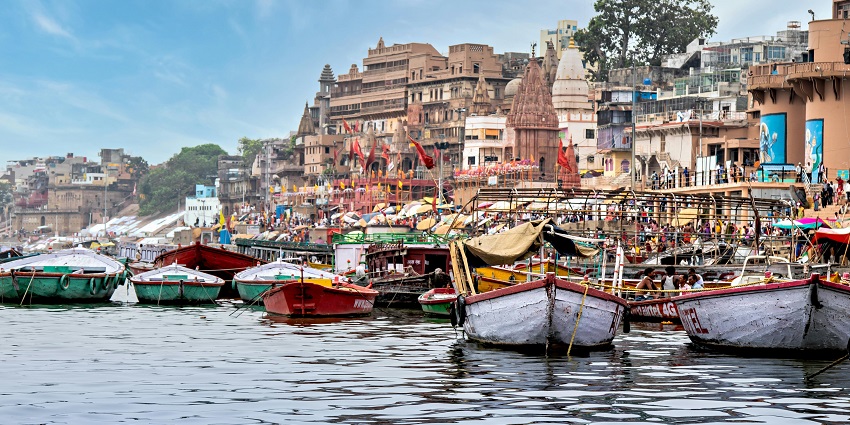 Varanasi ghat with boats docked along the calm river under a bright summer sky.