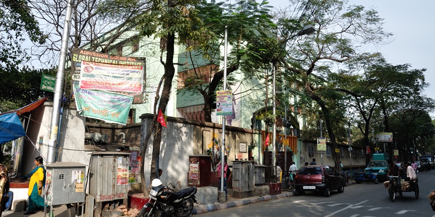 Street view with trees, parked vehicles, signboards, and people walking near a wall