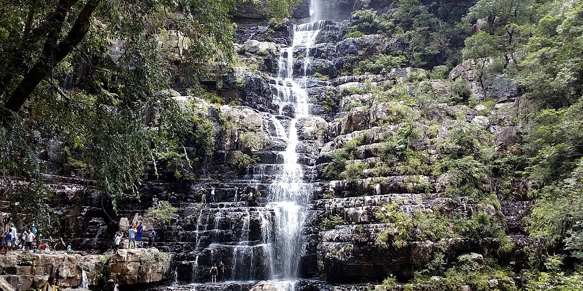 Multi-tiered waterfall cascading down rugged rock layers amidst dense forest greenery.