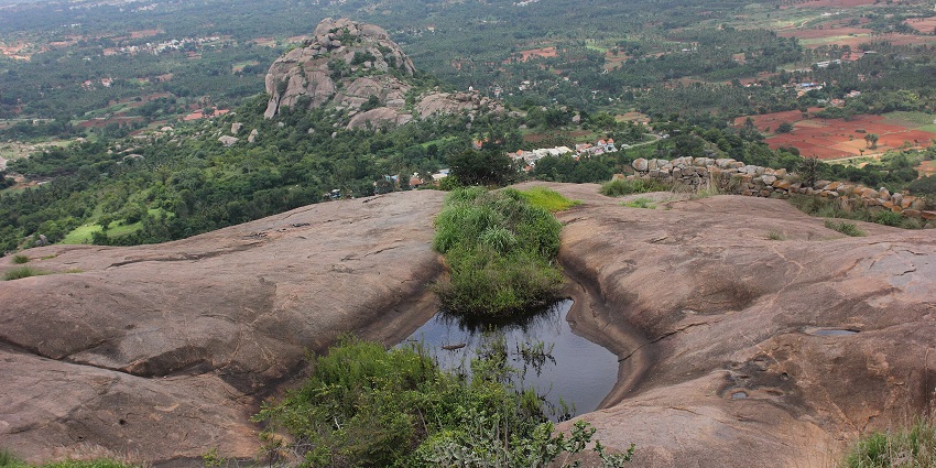 Rocky hilltop with a natural water pool, overlooking lush green valleys and distant hills.
