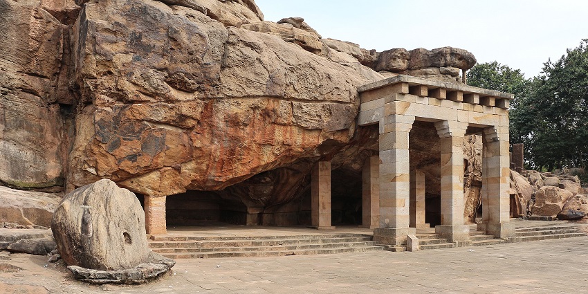 Entrance to the Udayagiri caves with three pillars, unique rock formations, and green trees.
