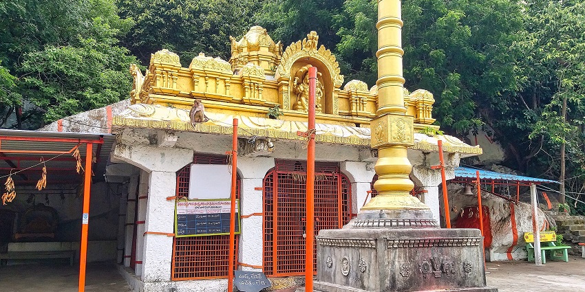 A small hillside temple with a golden tower, a stone pillar, and lush green forest around.
