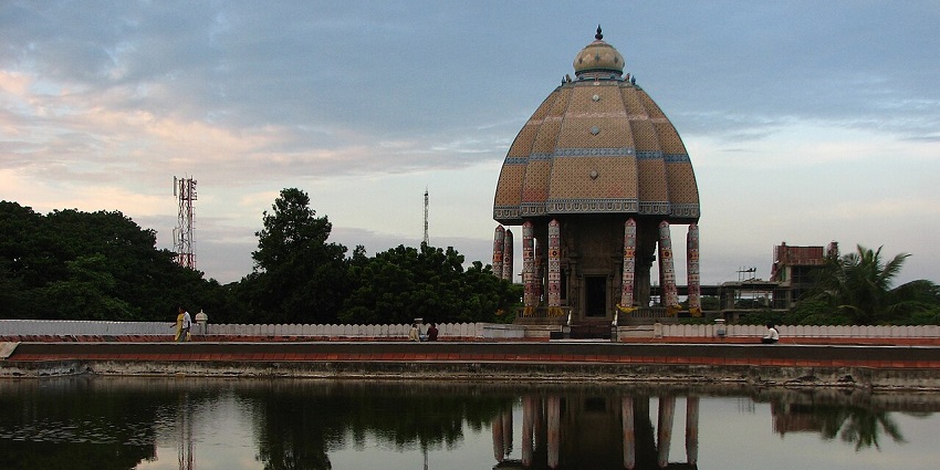 Reflection of Valluvar Kottam in a large water body in the front and distant greenery.
